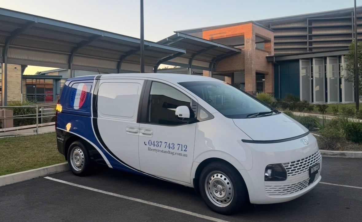 White and blue commercial van parked outside a modern building with a phone number displayed on its side