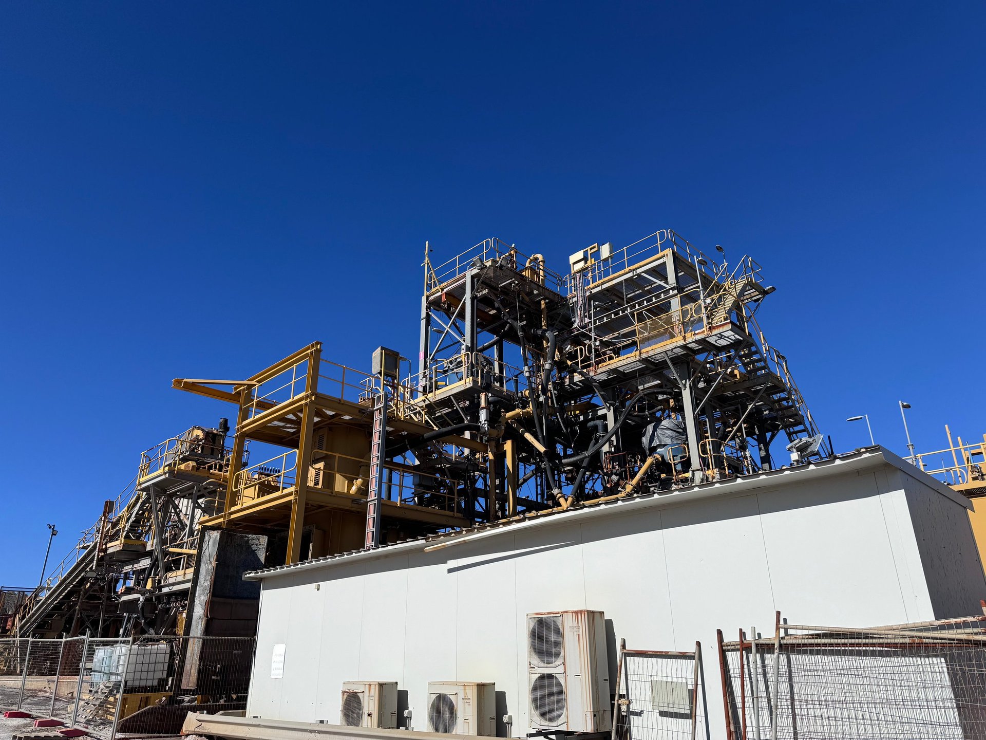 Large industrial mining or extraction facility with complex yellow metal structures and equipment against a clear blue sky