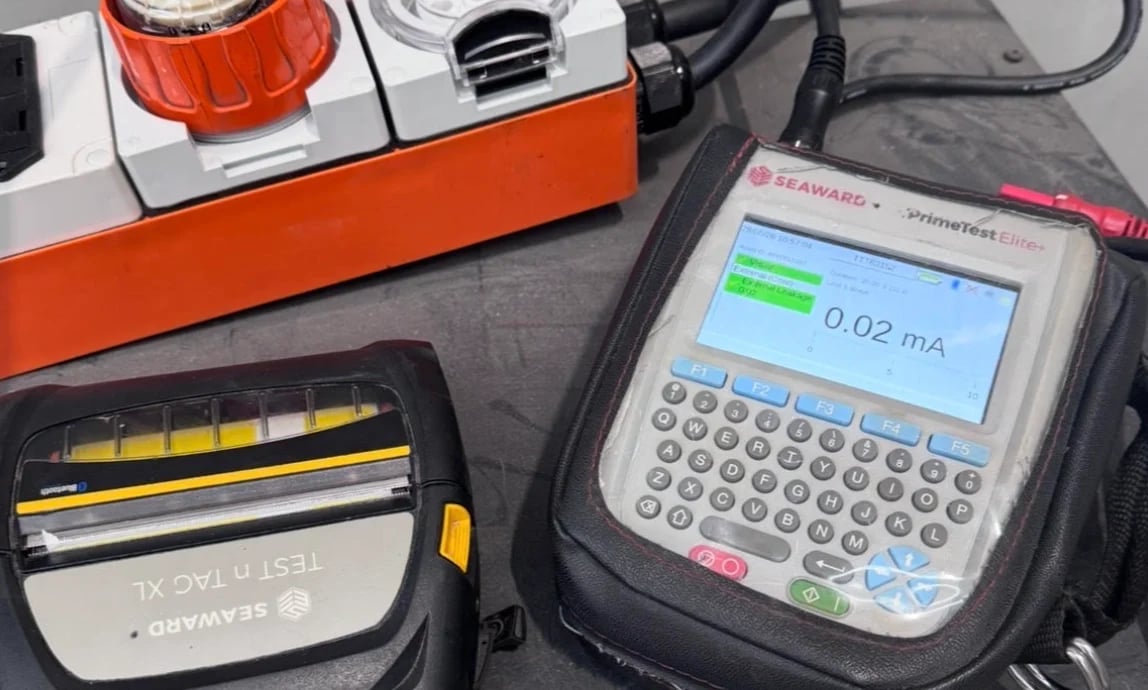 Electronic testing equipment including an orange and white control unit, handheld digital multimeter displaying 0.02 mA, and measurement tools on a gray surface