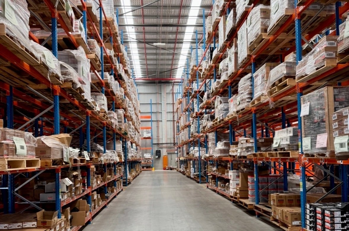 Interior of a large warehouse with rows of red and blue metal shelving units stacked with cardboard boxes and packages.