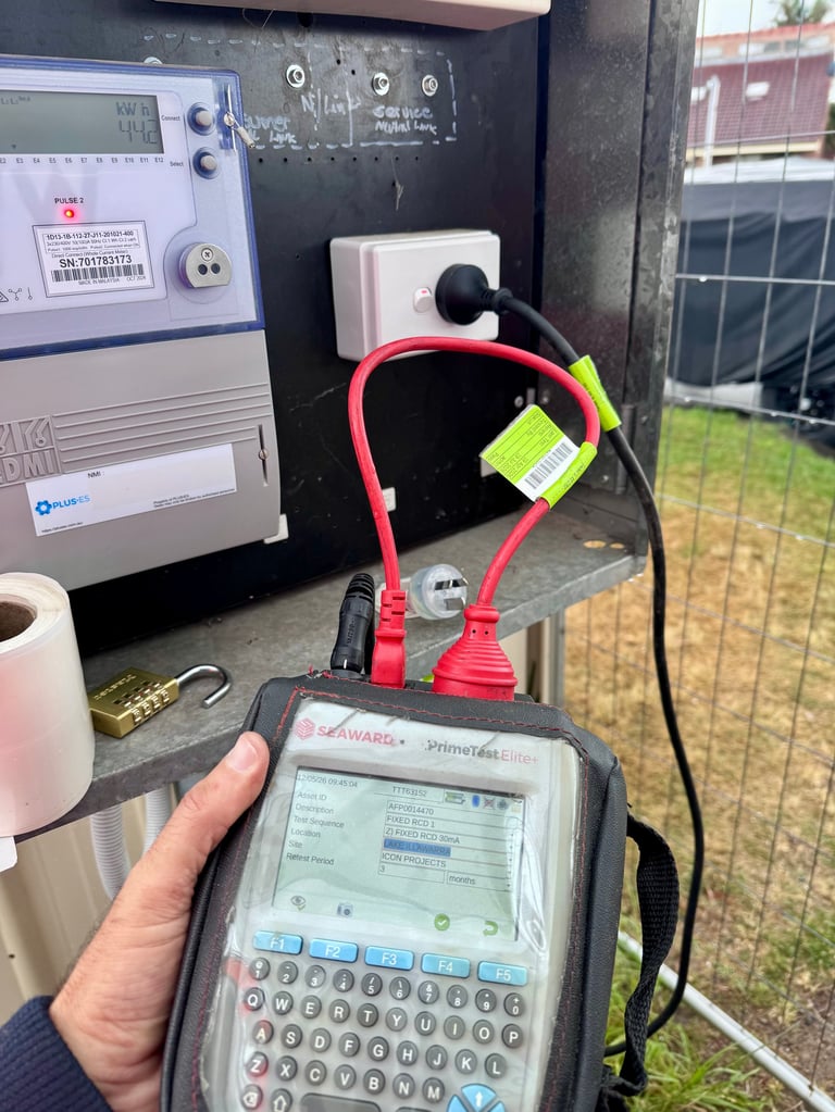 Person holding a portable test device with electrical equipment, red cable, and white outlet visible in the background