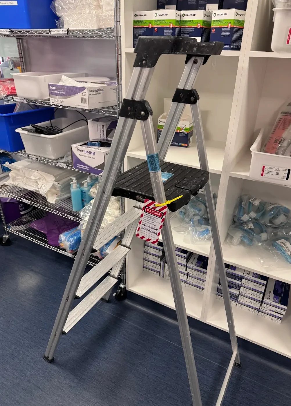 A tall aluminum step ladder with black platform and handles in a storage facility with shelves and organizational bins.