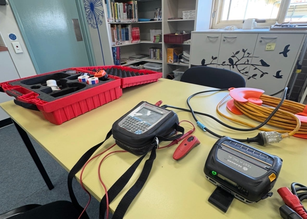 Classroom workbench with electronic testing equipment including multimeters, cables, and red storage bins