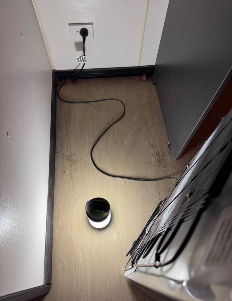 Overhead view of wooden floor with coiled black electrical cord, white outlet on wall, and white circular vent or fixture