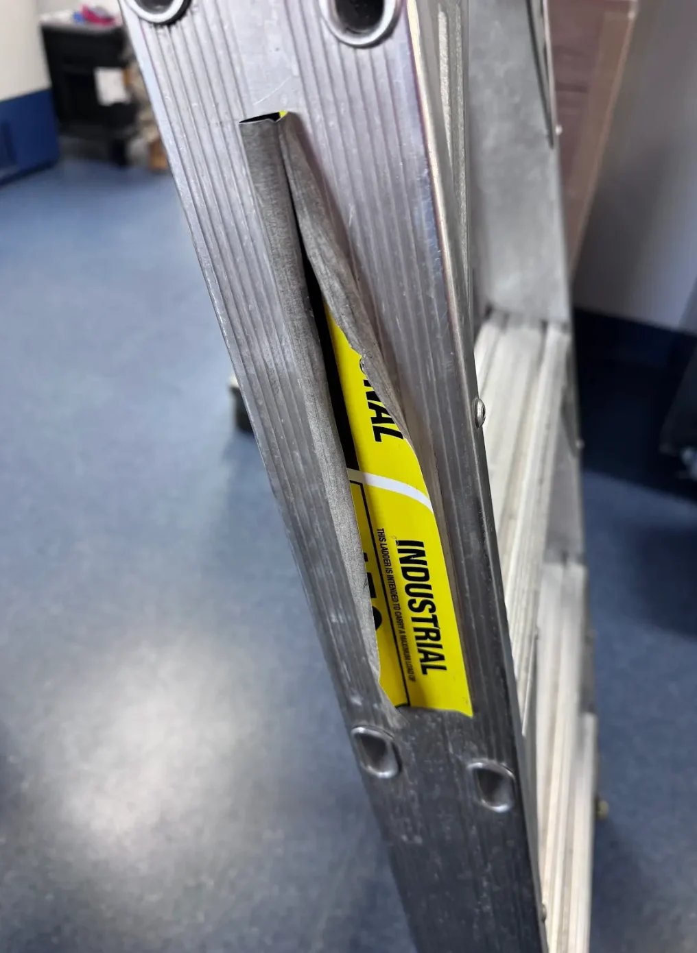 Close-up of an aluminum ladder with yellow label showing transmission warning text on blue flooring