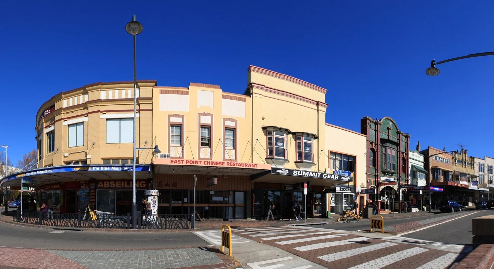 Historic cream-colored building with arched corner facade and storefront, street-level shops, clear blue sky, pedestrian crossing in foreground