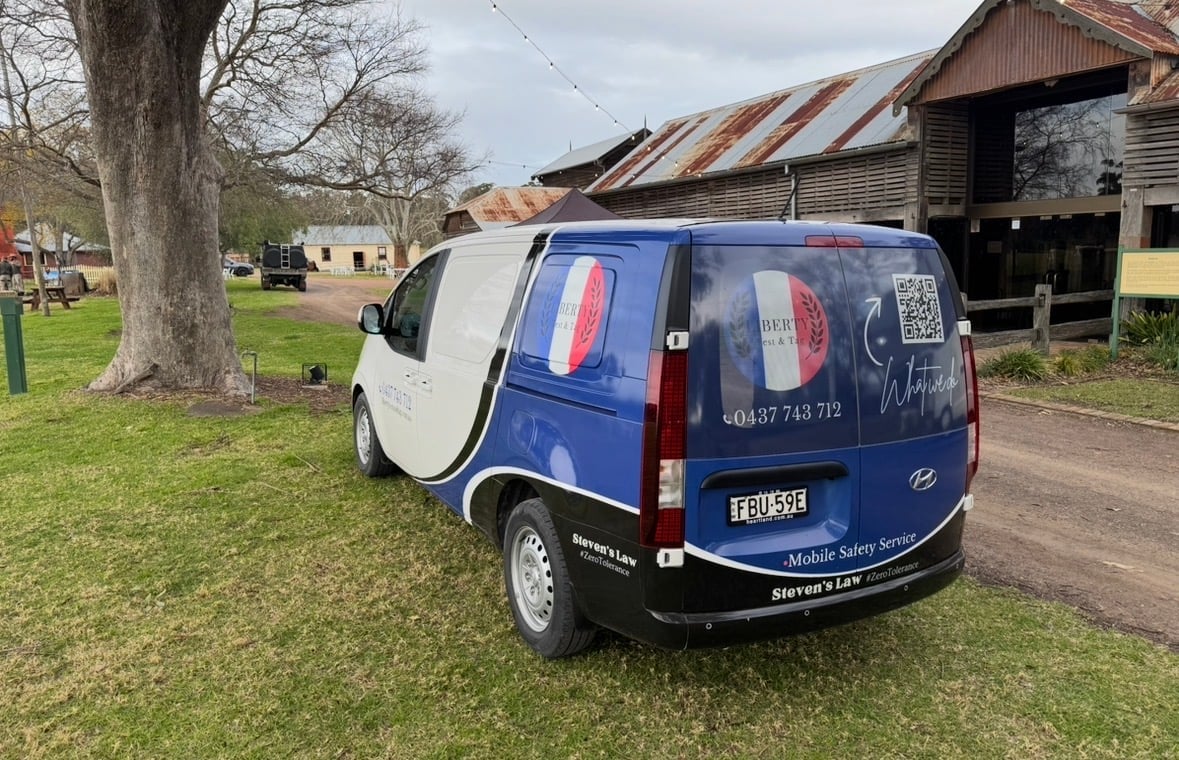 Blue and white electric van with open driver door parked on grass near rural buildings
