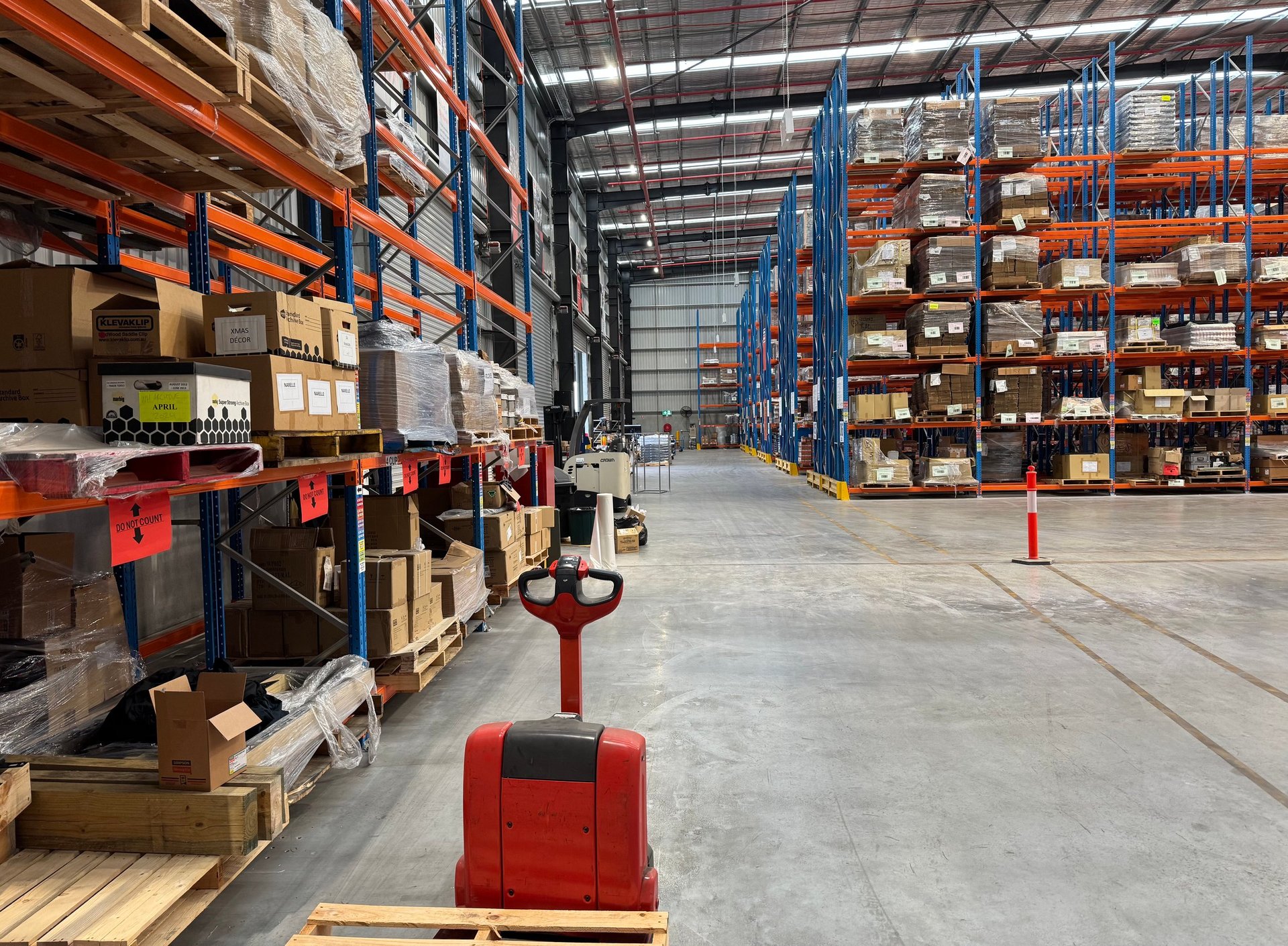 Large warehouse interior with tall metal shelving stacked with cardboard boxes, blue storage racks, and a red pallet jack in the foreground