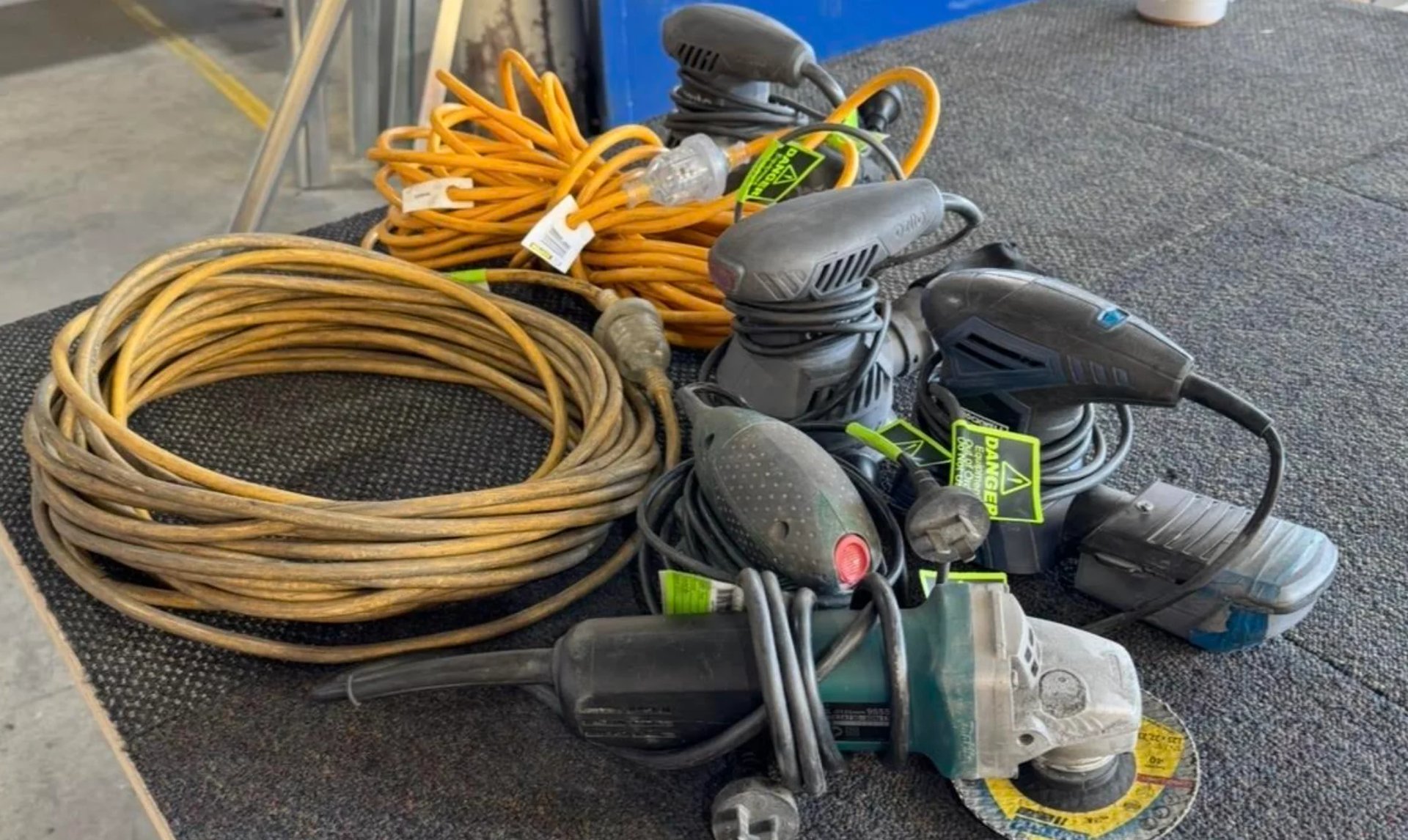 Collection of power tools and equipment including coiled yellow extension cord, black power drills, orbital sander, and various accessories on gray mat