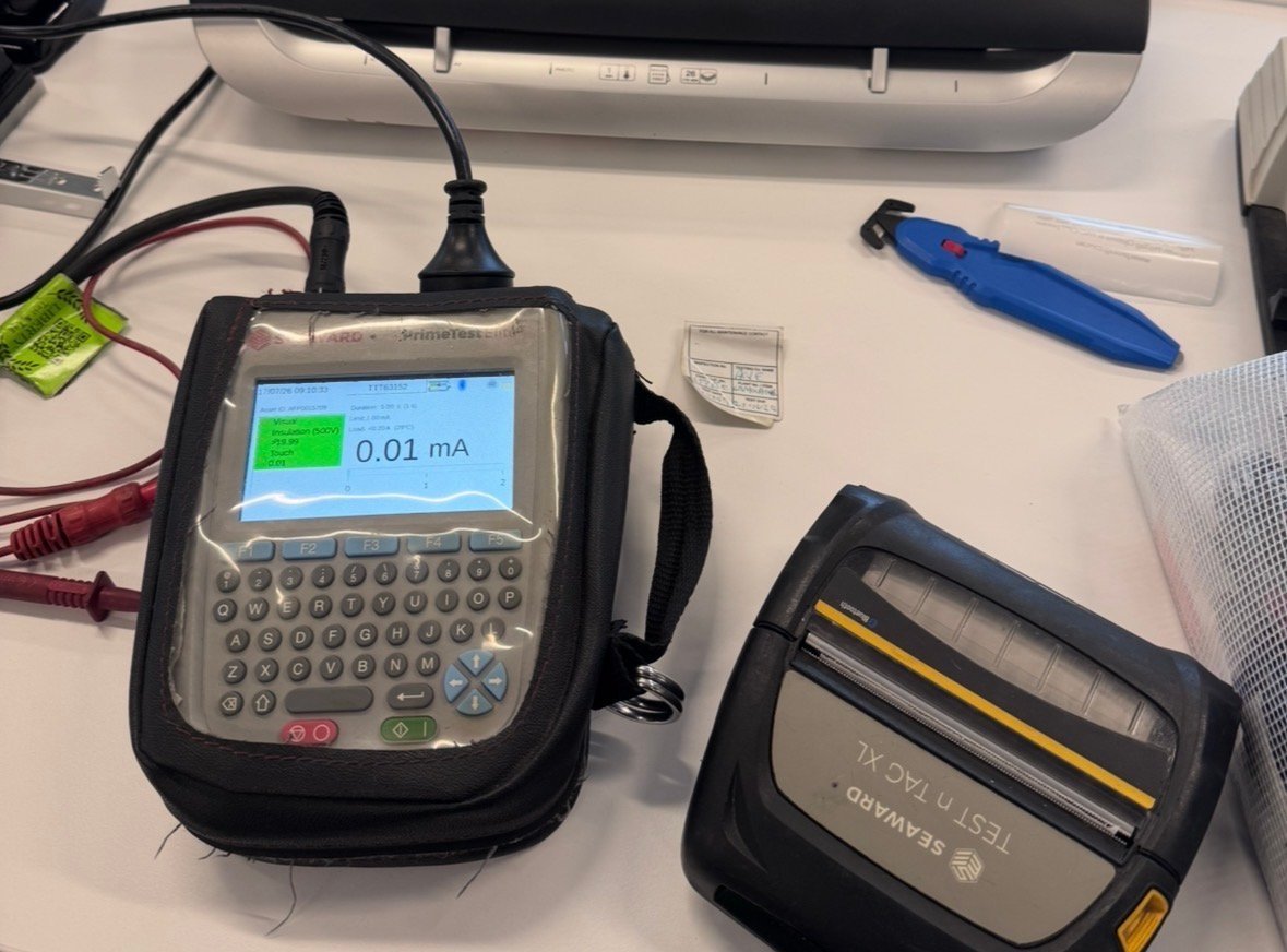 Digital handheld device with keyboard and green display showing 0.01 mA reading, with cables and accessories on white desk surface