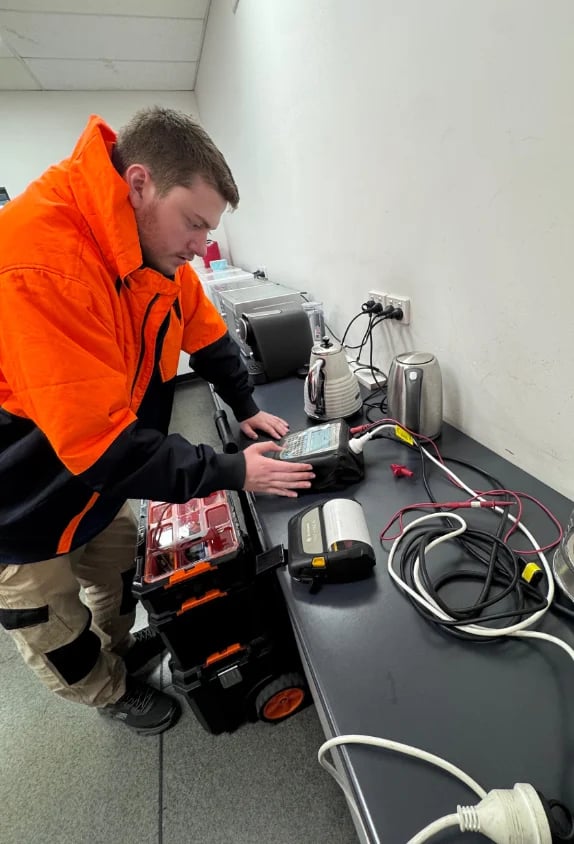 Technician in orange jacket inspecting electrical equipment and power tools on a workbench