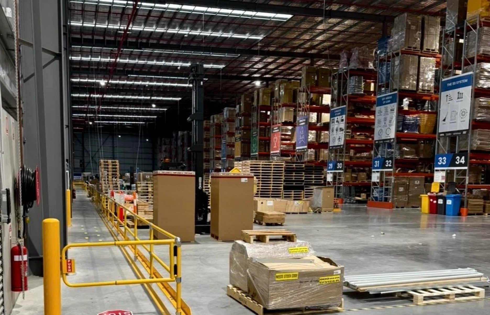Industrial warehouse interior with cardboard boxes stacked on pallets, yellow safety railing, and fluorescent ceiling lights