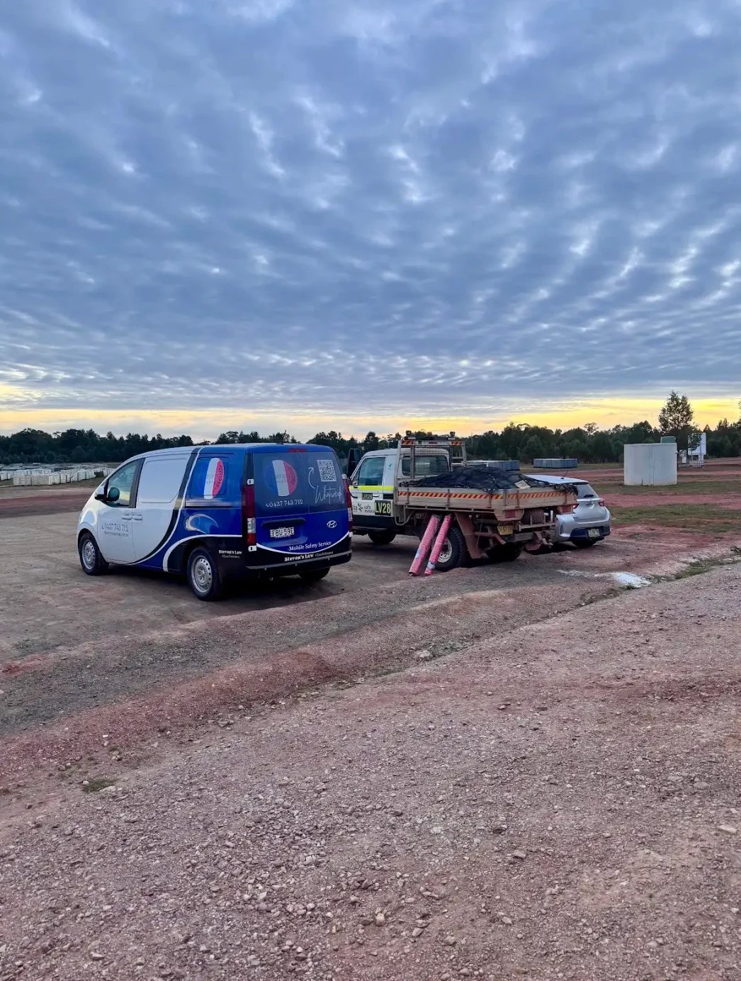 Two vans parked at a rural dirt lot with construction equipment and trees under a cloudy sky at dusk