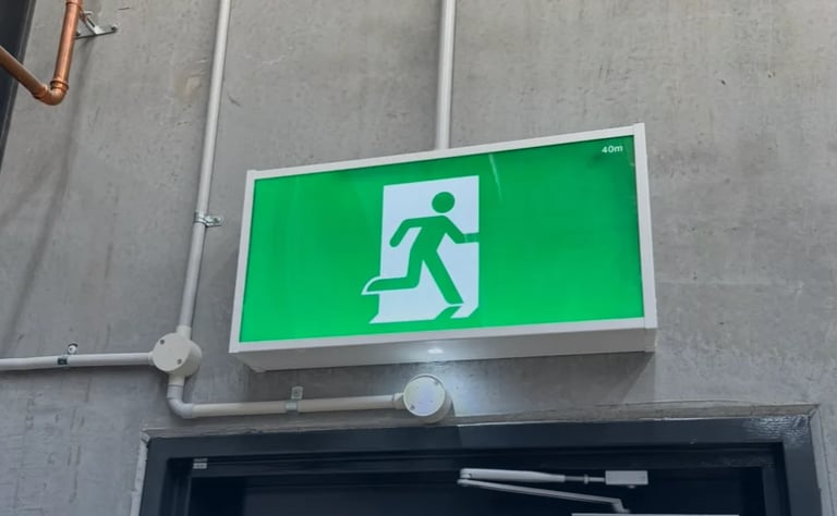 Illuminated green emergency exit sign with white running figure symbol mounted on gray wall above dark doorway