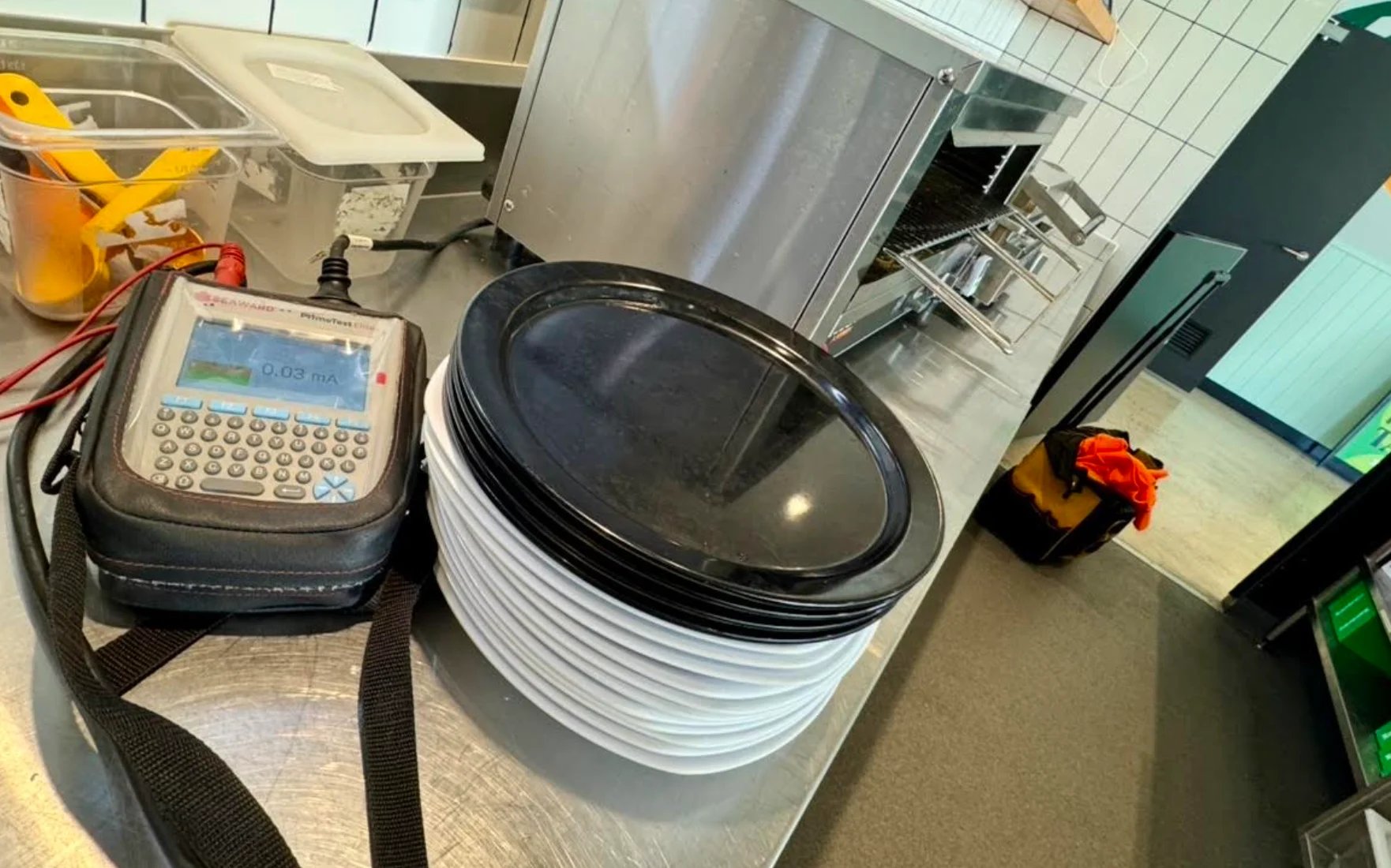 Kitchen counter with handheld digital device in carrying case, large white plastic container with black lid, and various tools on tiled surface