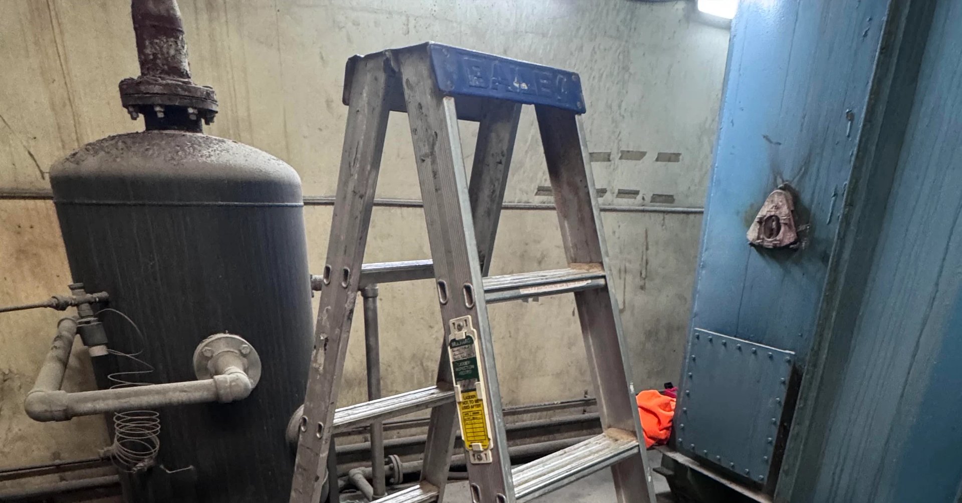 Industrial interior with metal tank on left, tall metal ladder in center, and blue door on right wall