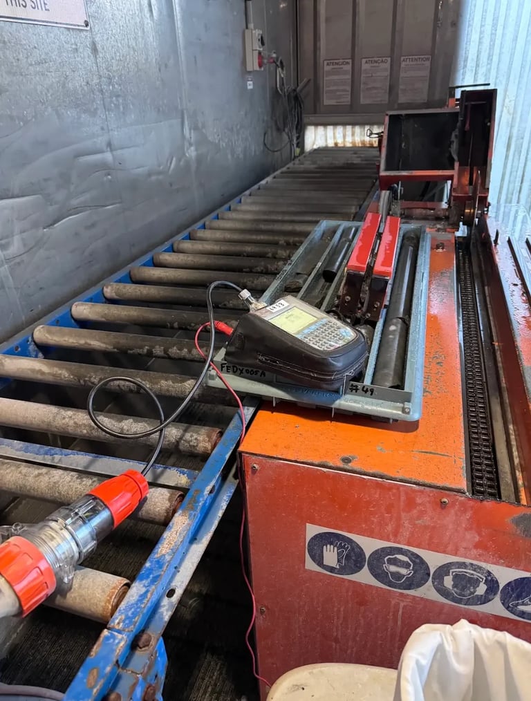 Industrial log processing machinery with conveyor rollers and red metal equipment inside a large warehouse or factory setting.