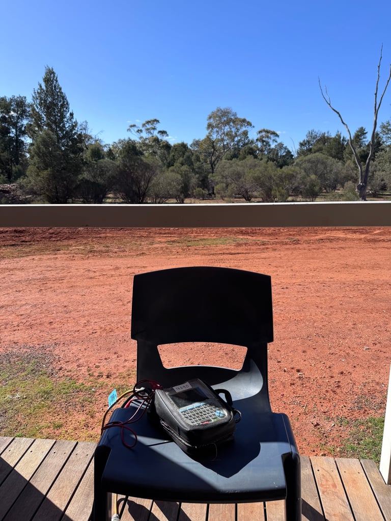 Seaward PrimeTest Elite+ portable appliance tester on a wooden deck, overlooking red earth field and eucalyptus trees under clear blue sky