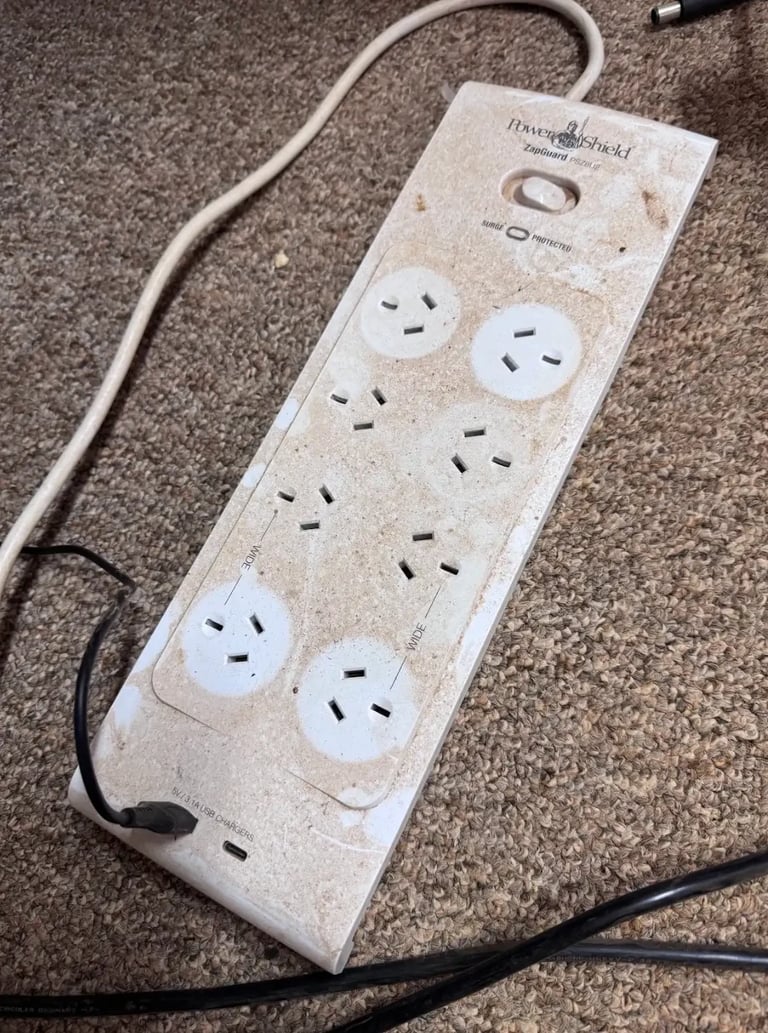 Beige power strip with multiple outlets and power cord on gray carpet, showing dust accumulation