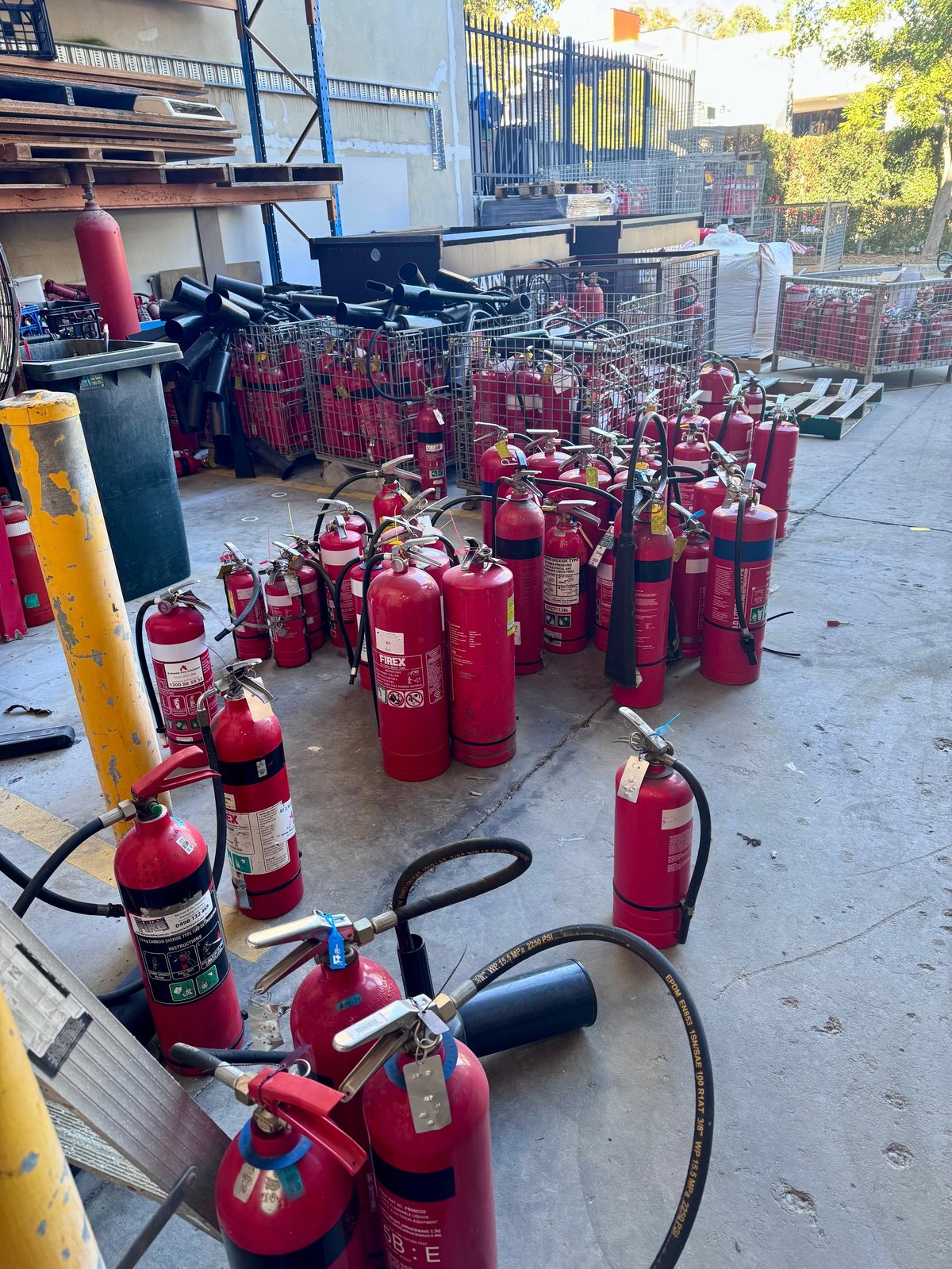 Multiple red fire extinguishers arranged on pavement outside industrial facility for inspection or maintenance