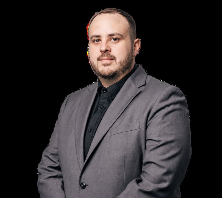 Professional headshot of a man with facial hair wearing a gray blazer and dark shirt against a black background