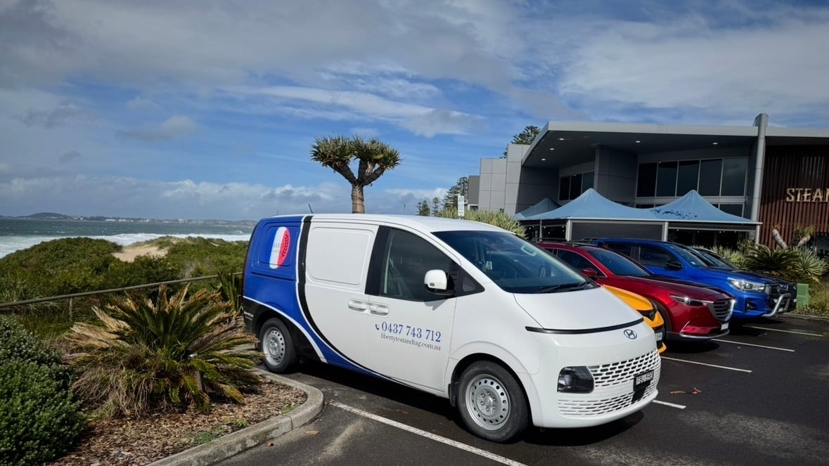 White and blue service van parked in coastal lot with red and blue vehicles and beach view in background