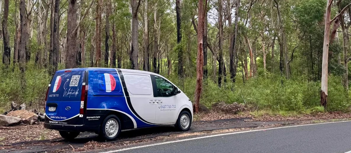 Blue and white commercial van parked on a paved road beside tall eucalyptus trees in a forest