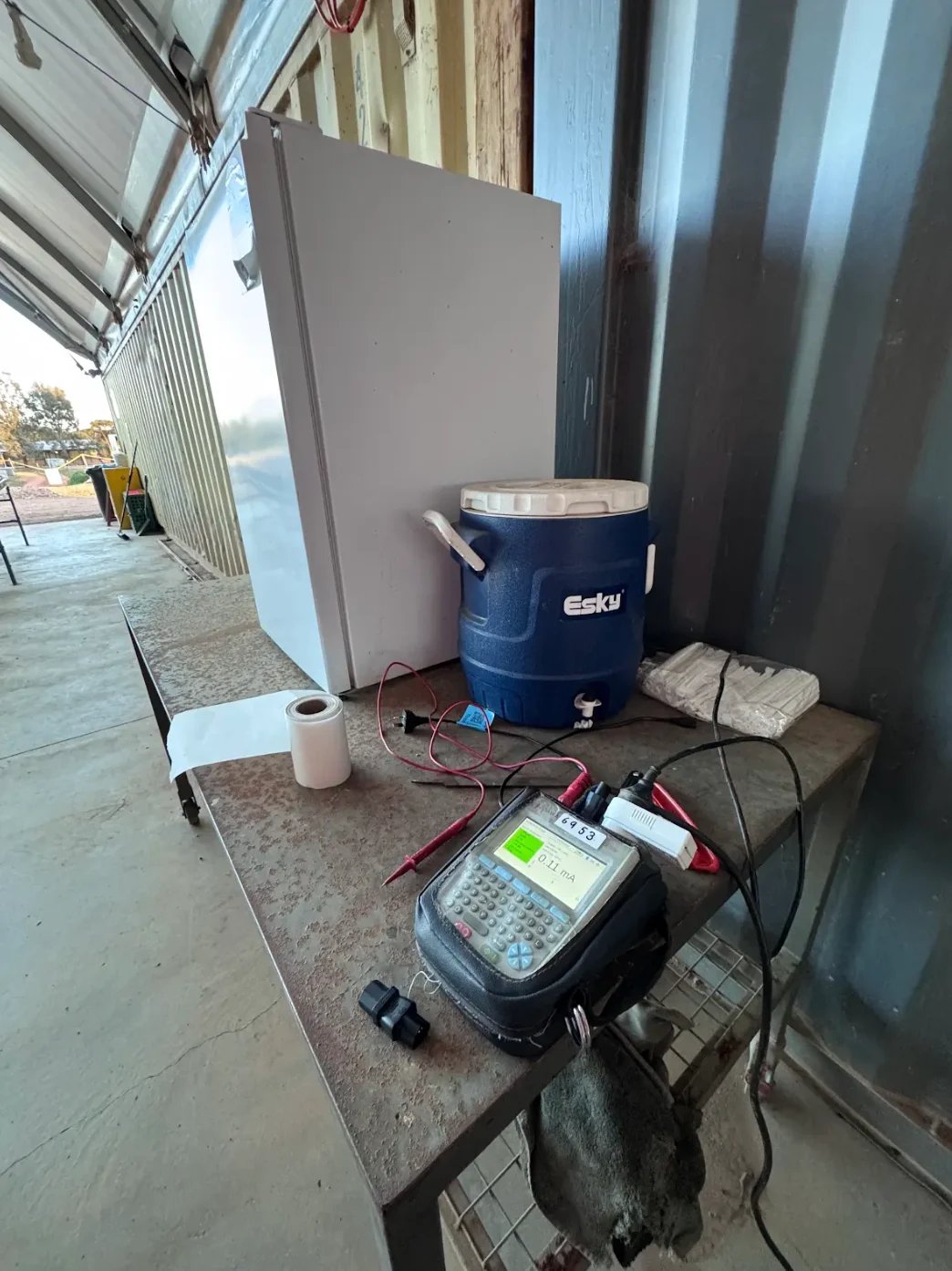 Laboratory equipment setup on a table including a white refrigerator, blue Esky cooler, and digital display device with electrical cables in an industrial warehouse space