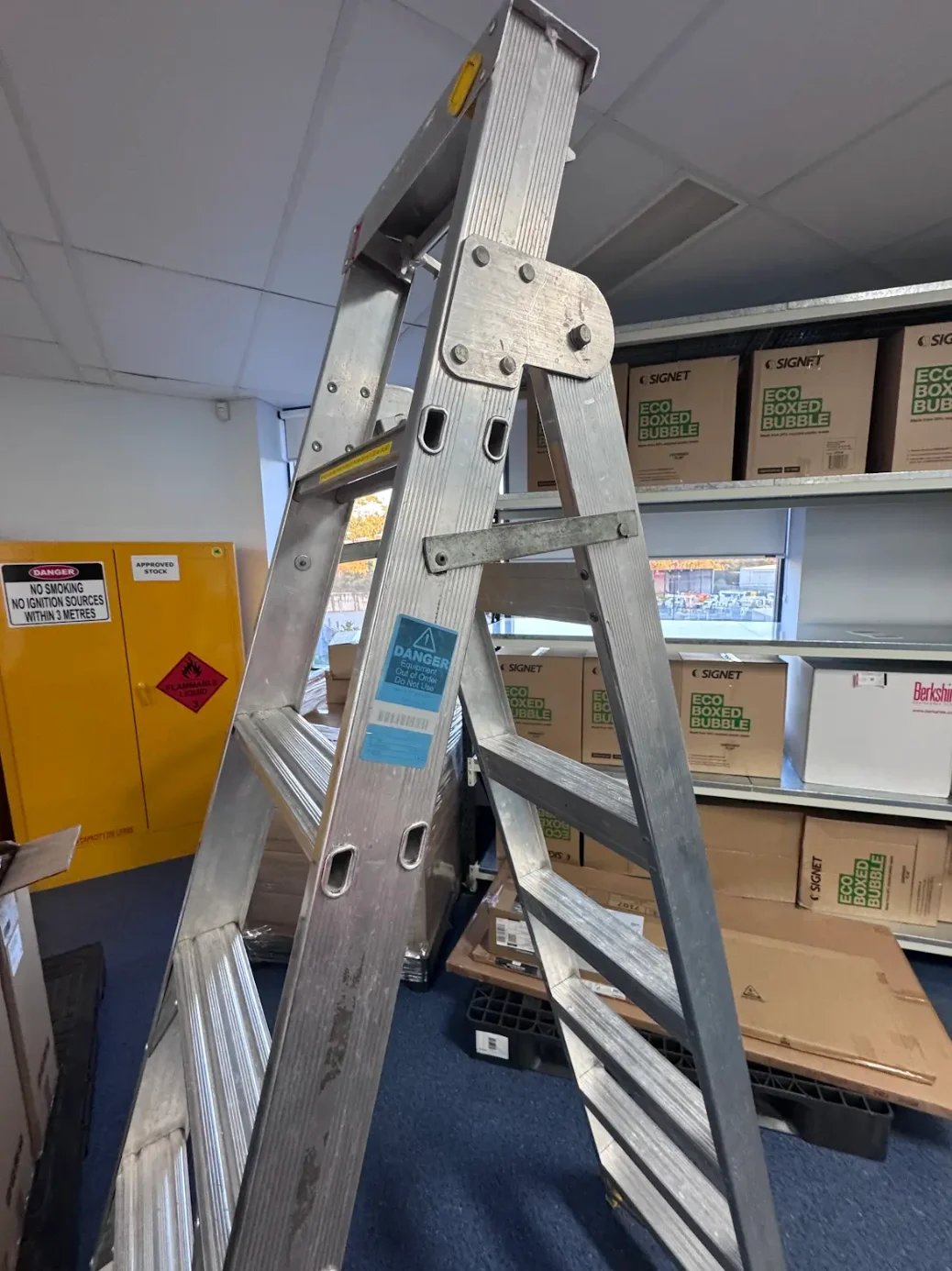 Aluminum step ladder positioned in an indoor storage area with cardboard boxes on shelves and a yellow cabinet in the background