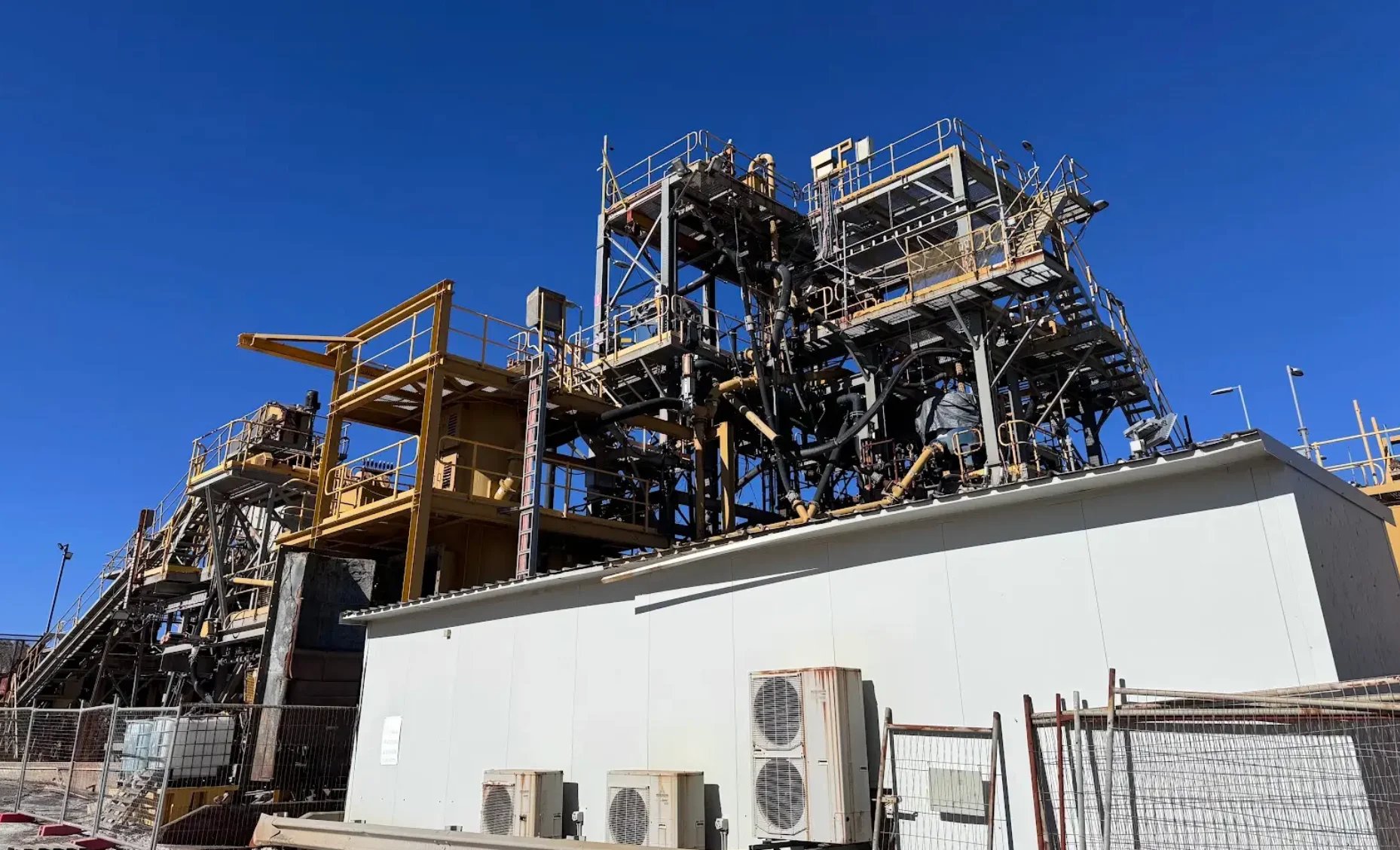 Industrial processing facility with large metal structures, yellow equipment, and white building with HVAC units against clear blue sky