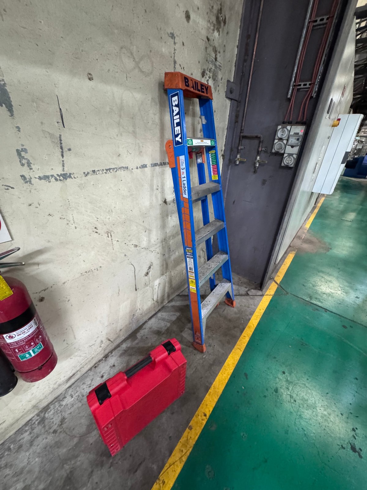Blue and orange ladder leaning against white concrete wall next to red safety barrier in an industrial garage setting