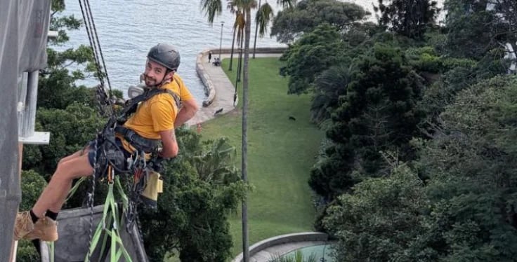 Person in yellow shirt rappelling down a tall pole with safety harness and helmet, palm trees and waterfront visible below
